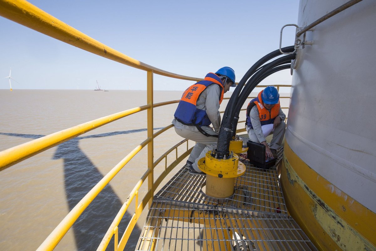 Two technicians in high-visibility gear inspect wind turbine components on an offshore platform.